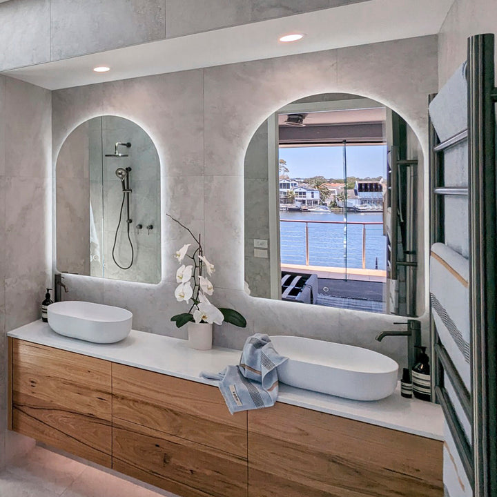 Lifestyle image: Two LED arch mirrors in a bathroom with two oval basins on stone top vanity, one mirror reflecting ocean.