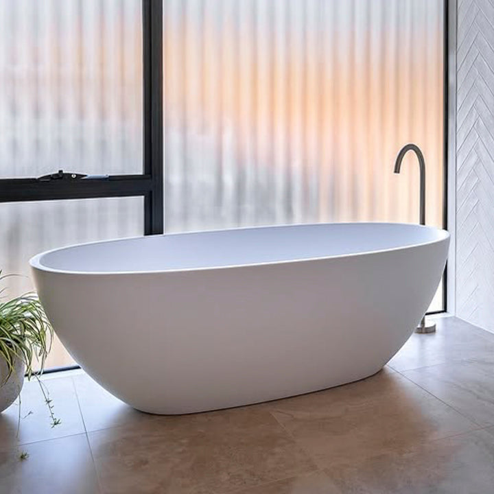 Byron Egg Oval Freestanding Bathtub in matte white with smooth curved design beside frosted floor-to-ceiling windows.