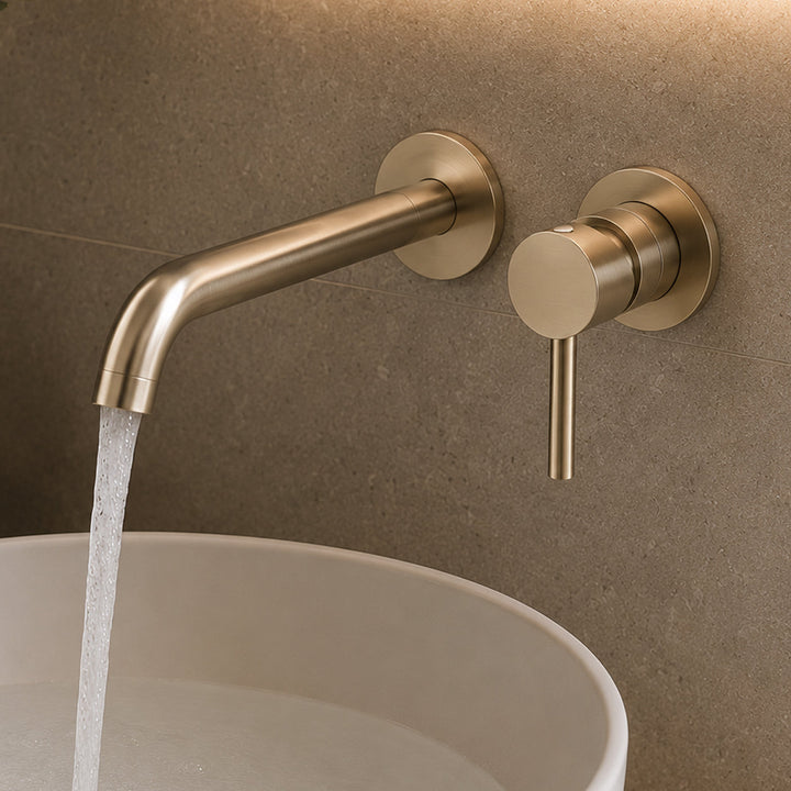 Brushed brass gold wall-mounted basin mixer with running water over a white round bathroom basin in a modern stone bathroom interior