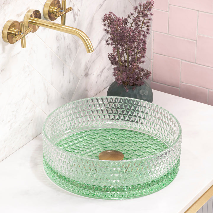 A lifestyle product shot featuring a green crystal glass basin on a stone countertop, accompanied by a brushed brass gold wall mixer matching the basin waste.