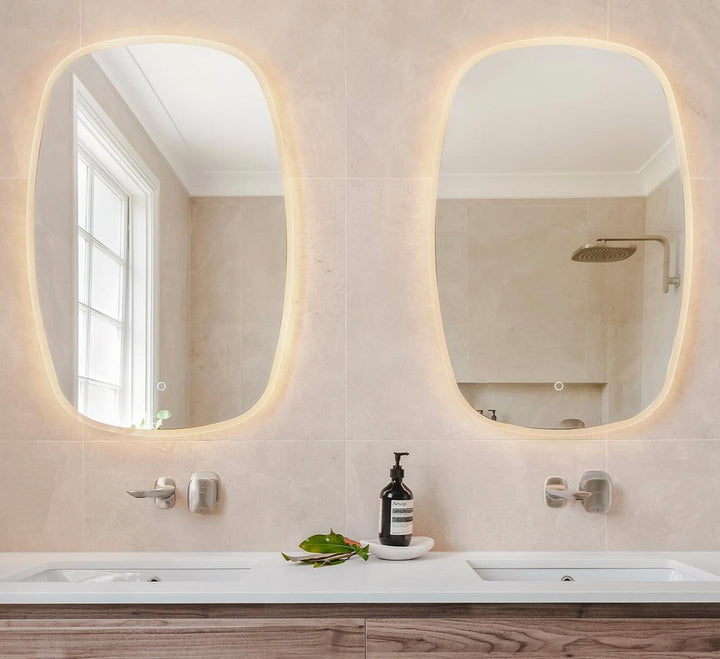 Double oblong LED mirrors with warm backlighting above dual sinks in a modern bathroom with beige tiles and wood vanity.