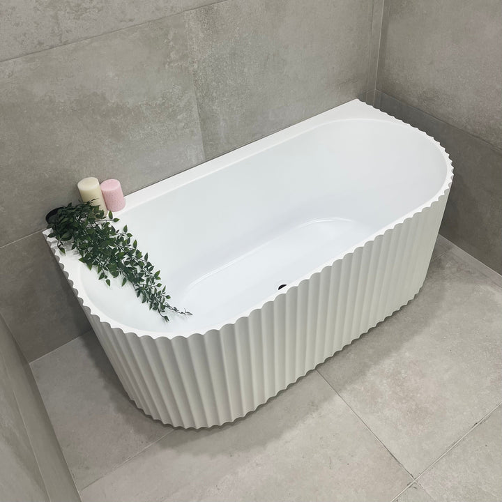 Back to wall freestanding bathtub in matte white with fluted exterior, positioned against tiled wall with bath accessories on edge